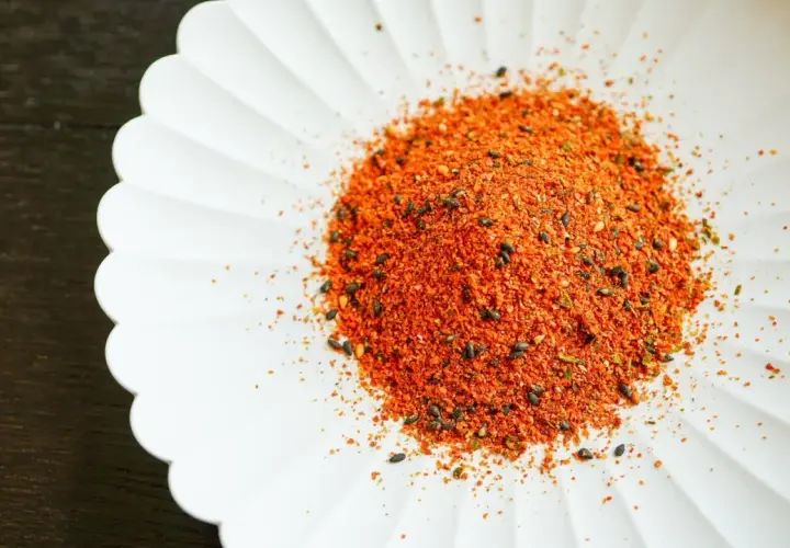 A pile of vibrant Japanese 7 spice blend, featuring chili flakes and black sesame seeds, sits on a white, fluted dish.