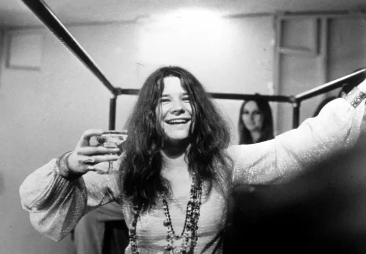 Janis Joplin, z kieliszkiem w dłoni, uśmiecha się szeroko. Jej charakterystyczne włosy opadają na ramiona, a na szyi wisi naszyjnik. W tle widać inną kobietę. To zdjęcie przypomina o jej niezapomnianych **janis joplin utworach**.