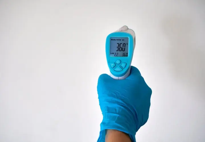 Ręka w niebieskiej rękawiczce trzyma termometr bezdotykowy, pokazujący 36.0°C. Idealne miejsce, gdzie mierzyć temperaturę na czole lub skroni.