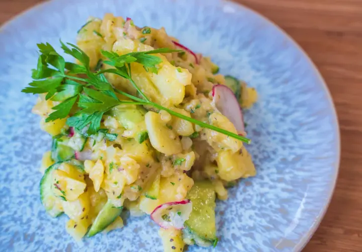 Ein köstlicher Kartoffelsalat mit Gurken, Radieschen und frischer Petersilie auf einem blauen Teller.