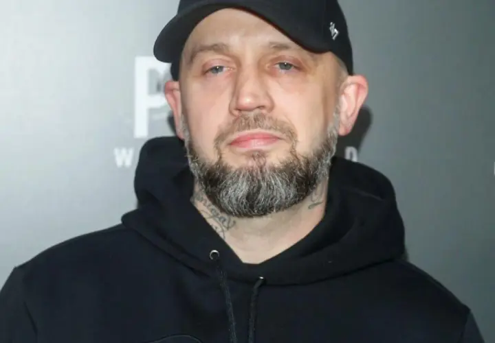Mężczyzna w czarnej bluzie z kapturem i czapce z daszkiem, z brodą i tatuażami na szyi. Pikers ile ma lat?