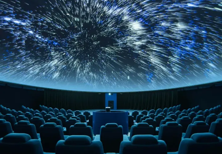 Wnętrze planetarium, gdzie na kopule wyświetlane są rozgwieżdżone niebo i galaktyki. Widownia z niebieskimi fotelami czeka na seans.