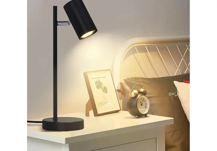 Lampara mesilla negra ilumina un cuadro y un despertador vintage en una mesita de noche junto a una cama.