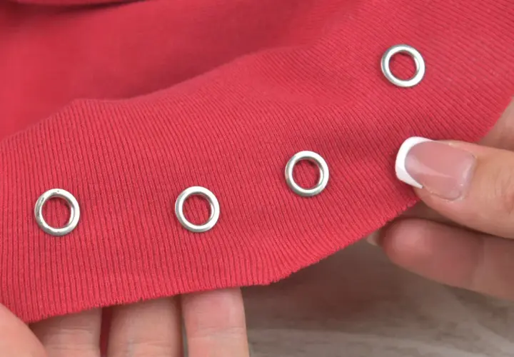 Demonstration of how to put metal rings in fabric. A hand holds red fabric with several silver grommets already attached, and another grommet is about to be placed.