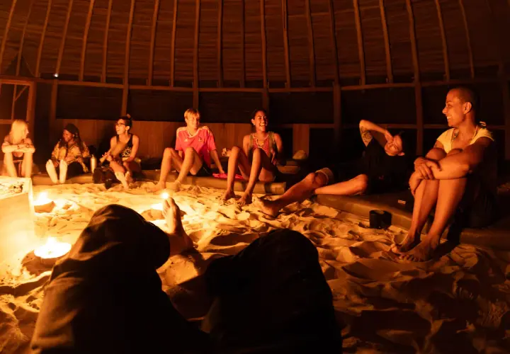 Grupa ludzi siedzi na piasku w okrągłej, drewnianej chacie, otoczona świecami. Rozmawiają i śmieją się, przygotowując się do ceremonii. Ayahuasca cena może być różna, ale to doświadczenie jest bezcenne.