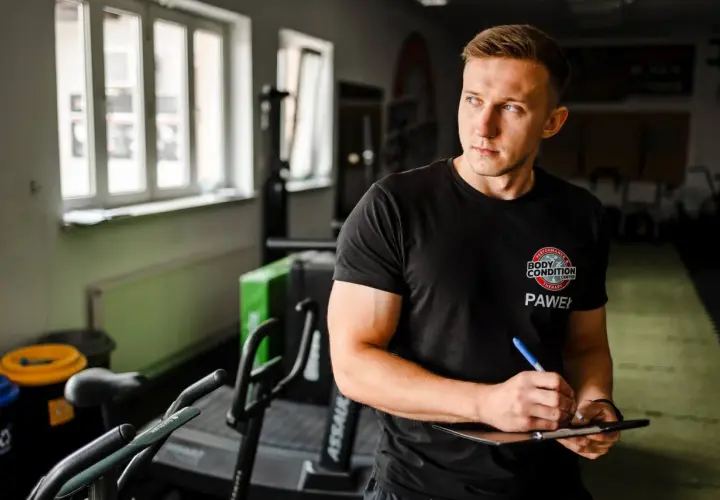 Trener Paweł tworzy spersonalizowany plan treningowy na biceps, notując ćwiczenia na tablicy.