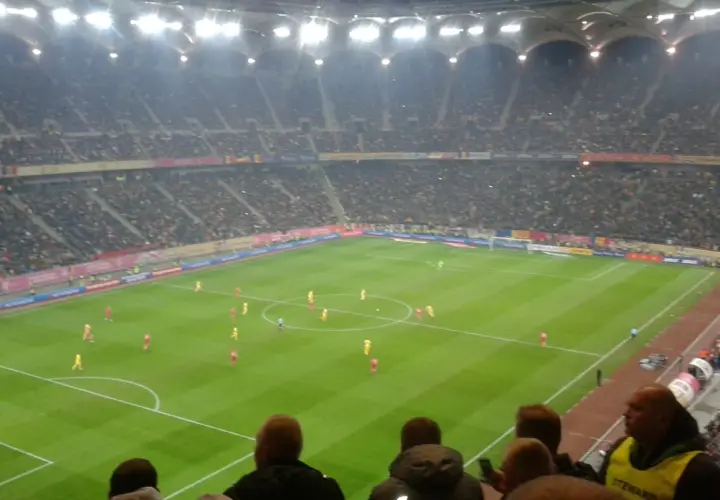 Mecz piłki nożnej na stadionie, tłumy kibiców, flagi Polski i Rumunii.