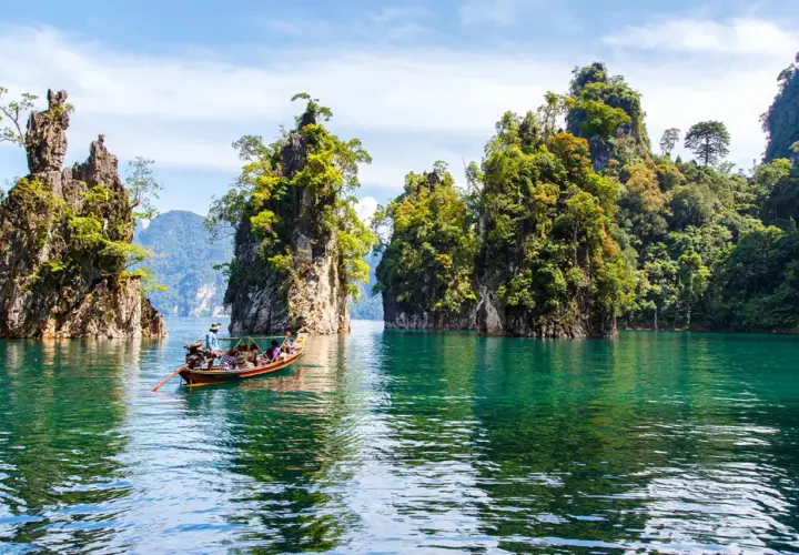 Bootfahrt durch smaragdgrünes Wasser im **Thailand Regenwald**, vorbei an steilen, bewachsenen Felsen unter blauem Himmel.