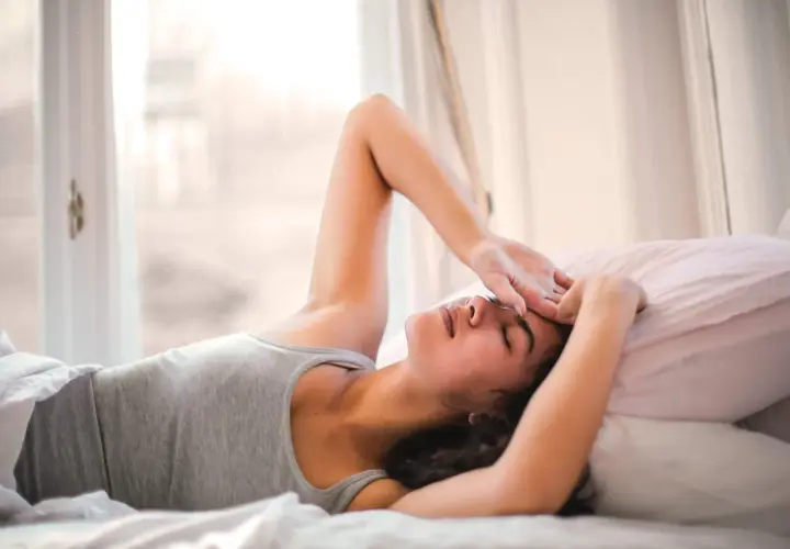 Ragazza sdraiata a letto, con la mano sulla fronte, sembra che non riesca a svegliarsi la mattina. Luce soffusa dalla finestra.