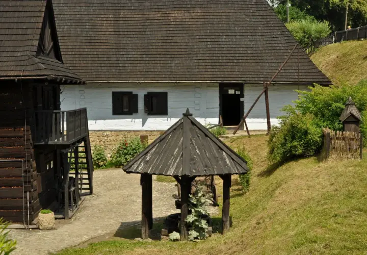 Skansen w Dobczycach: drewniane chaty, studnia i zielone wzgórza tworzą sielski krajobraz.