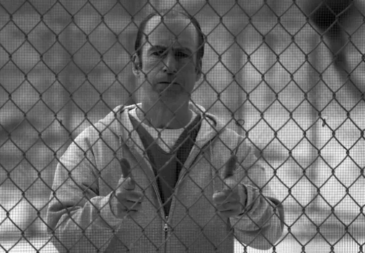 Un uomo dietro una rete metallica, con un'espressione pensierosa. Il suo gesto con i pollici in su suggerisce un "tutto bene" che contrasta con la sua situazione, evocando il **better call saul significato** di apparenza ingannevole.