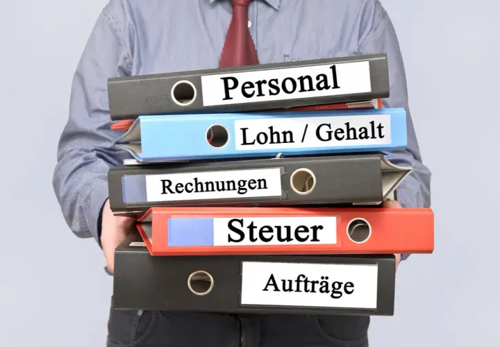 Ein Mann hält einen Stapel Ordner mit den Beschriftungen "Personal", "Lohn / Gehalt", "Rechnungen", "Steuer" und "Aufträge". Die Ordner stapeln sich und deuten auf die Komplexität der Verwaltung des zu versteuernden Einkommens hin.