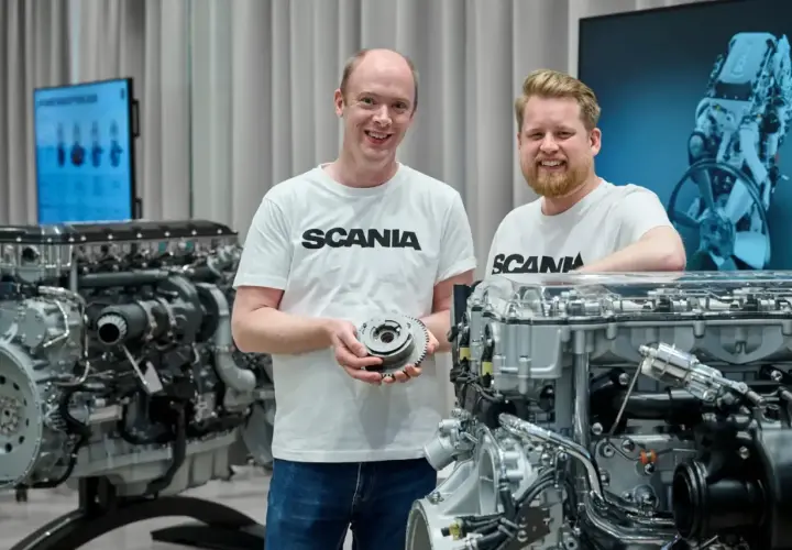 Dwóch mężczyzn w koszulkach Scania prezentuje części silnika. W tle widać potężny silnik Scania V8.