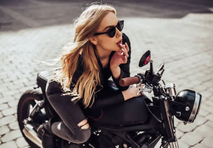 Chica rubia con gafas de sol y un perfecto outfit motero mujer, apoyada en una moto clásica.