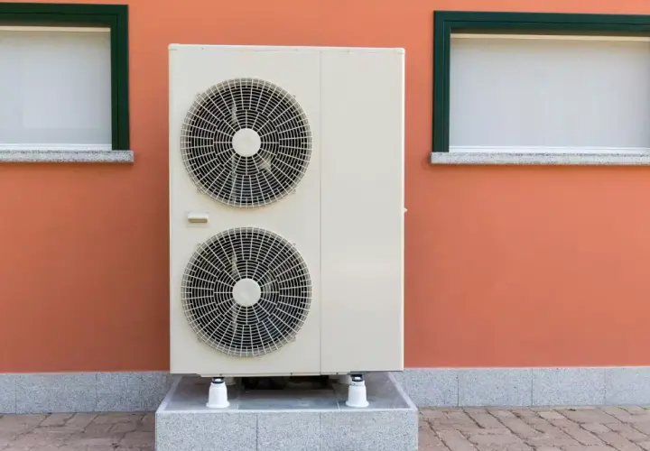 Unidad exterior de bomba de calor con dos ventiladores, instalada junto a una pared naranja con dos ventanas.