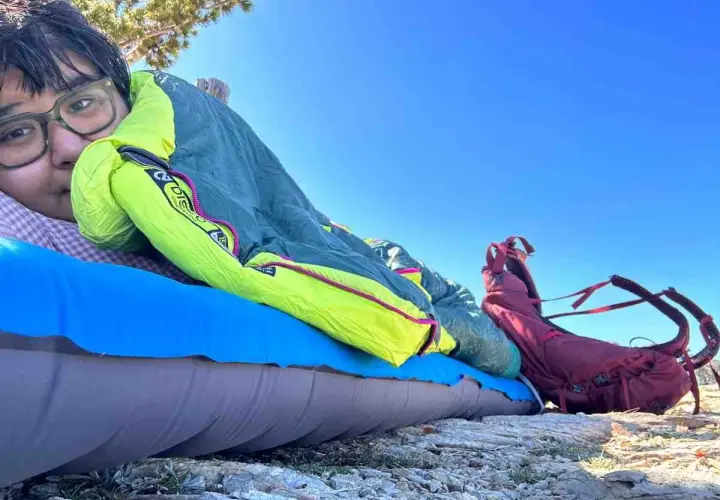 A person rests on a sleeping pad under a bright blue sky, bundled in a Nemo Disco 15 weight sleeping bag. A red backpack sits nearby.