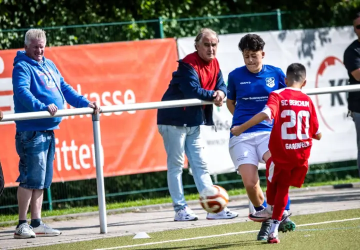 Fußball-Eltern schauen ihren Kindern beim Spiel zu. Ein Junge im roten Trikot mit der Nummer 20 spielt den Ball.