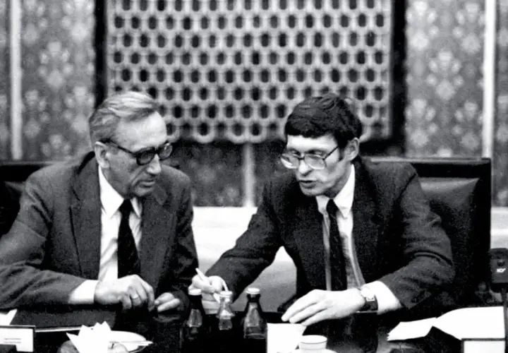 Dwóch mężczyzn w garniturach przy stole, dyskutujących o historii politycznej Polski. Antoni Dudek analizuje dokumenty.