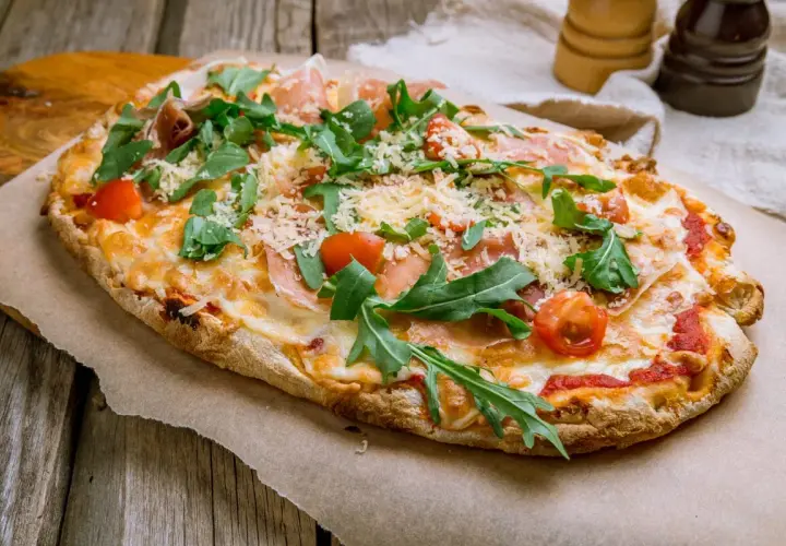 Pyszna pinsa romana z rukolą, pomidorkami i szynką parmeńską, posypana parmezanem, na drewnianym stole.