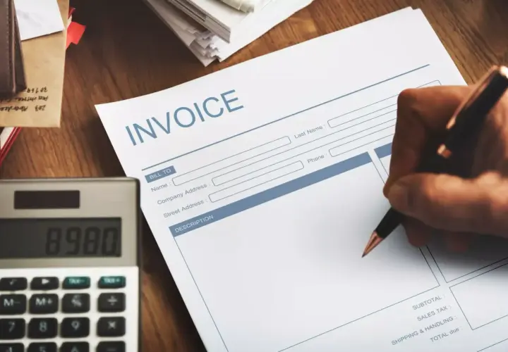 Ręka pisze na fakturze "INVOICE". Obok leży kalkulator. Faktura VAT marża - co oznacza dla kupującego?