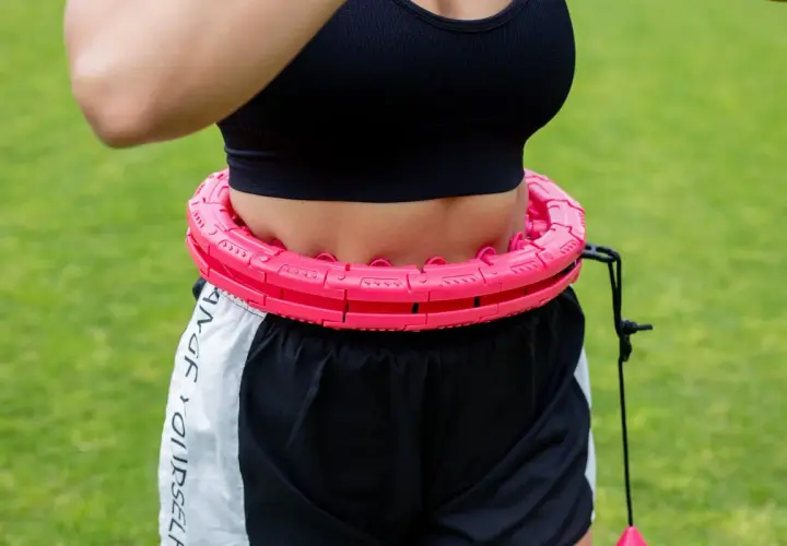 Kobieta w sportowym topie i szortach wykonuje ćwiczenia z hula hop na trawie. Różowe, segmentowe hula hop z obciążnikiem.