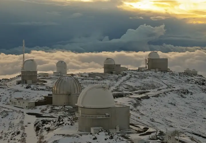 Obserwatoria astronomiczne na ośnieżonym szczycie, otoczone morzem chmur. W oddali widać złote światło zachodzącego słońca, a na pierwszym planie "la silla" - jedno z kopułowych budynków.
