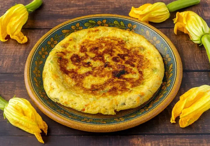 Frittata dorata con fiori di zucca, servita su un piatto decorato, circondata da fiori freschi.
