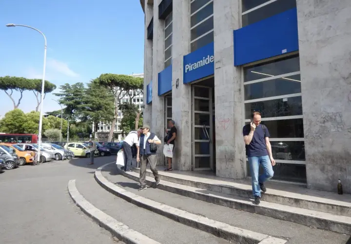 Uscita della metro Piramide a Roma Ostiense. Persone entrano ed escono dall'edificio, mentre auto sono parcheggiate nelle vicinanze.