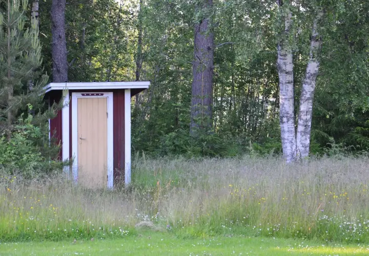 Rött utedass med gul dörr, omgivet av högt gräs och träd. Här gäller utedass regler för en trevlig vistelse.