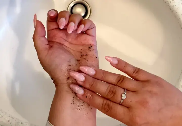 Exfoliante de café en la mano para quitar el autobronceador. Uñas largas y rosadas, anillo de corazón.