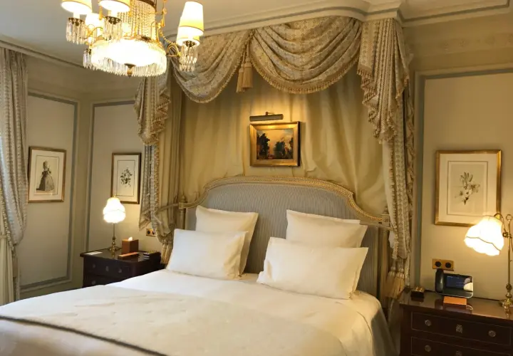 Elegancka sypialnia w hotelu Ritz w Paryżu, z baldachimem nad łóżkiem, kryształowym żyrandolem i zabytkowymi obrazami.