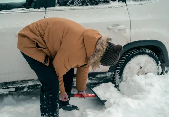 Osoba w zimowej kurtce odśnieża samochód uwięziony w zaspie śnieżnej.