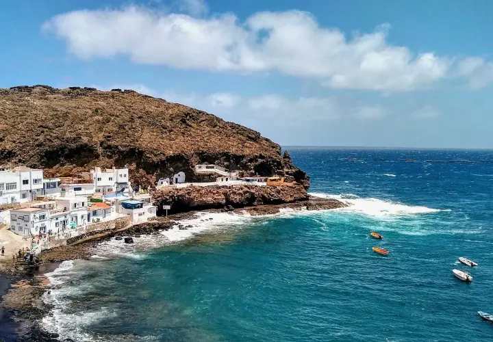Pueblo costero blanco anidado en la ladera rocosa de **Tufia**. El mar azul turquesa choca contra las rocas, con pequeñas embarcaciones flotando.