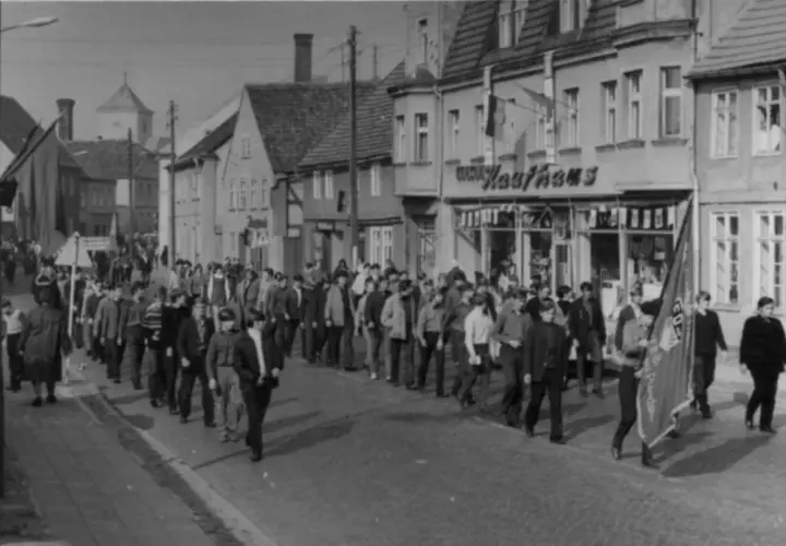 Demonstration am 7. Oktober, Tag der Republik DDR. Menschen marschieren durch eine Straße mit Geschäften, Fahnen wehen.