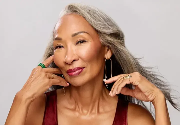 Mujer con cabello gris, con un elegante efecto de grey blending, sonríe con labios rojos y joyas doradas y verdes.