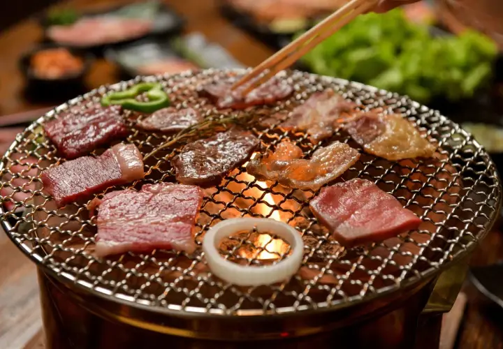 Enjoying delicious yakiniku at home. Slices of raw beef sizzle on a grill over glowing coals, with green peppers and an onion ring adding color.