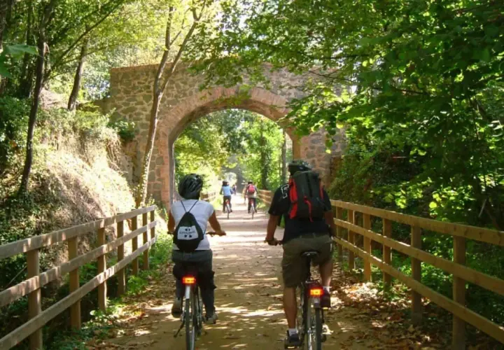 Ciclistas disfrutan de la vía verde Olot Girona, pedaleando bajo un antiguo arco de piedra rodeado de exuberante vegetación.