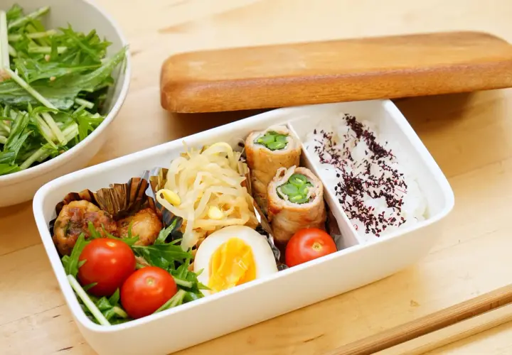 A delicious rice bento box with pork rolls, chicken meatballs, a hard-boiled egg, cherry tomatoes, and a side salad.
