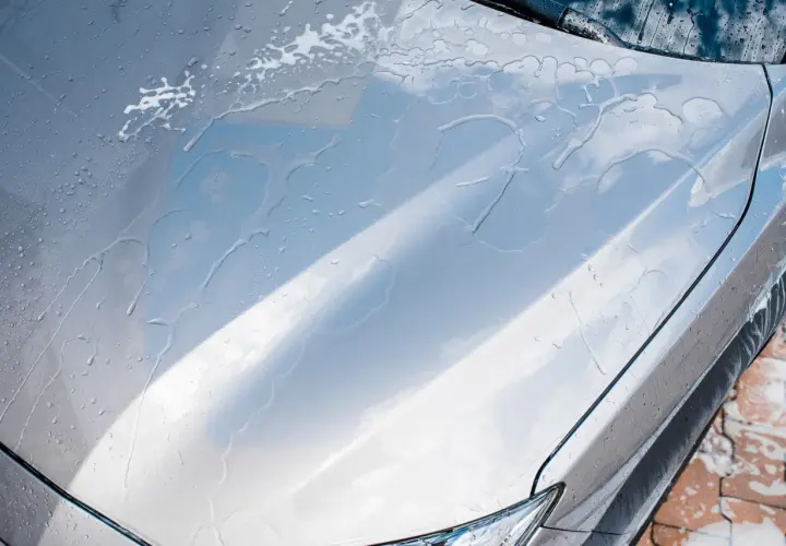 Krople wody spływają po masce srebrnego auta, tworząc smugi. Czy po woskowaniu płukać auto? Tak, aby uzyskać idealny połysk.