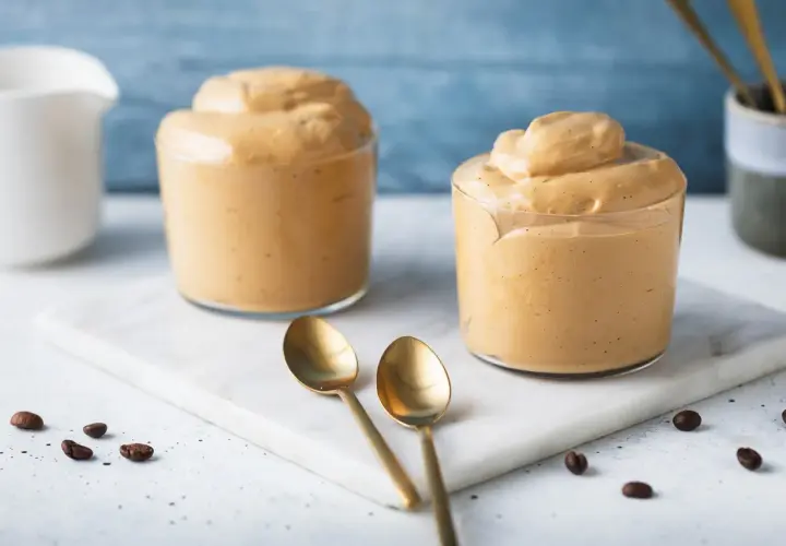 Due bicchieri di cremosa schiuma di caffè, perfetta per la tua ricetta crema di caffè senza panna. Due cucchiaini dorati e chicchi di caffè completano la scena.