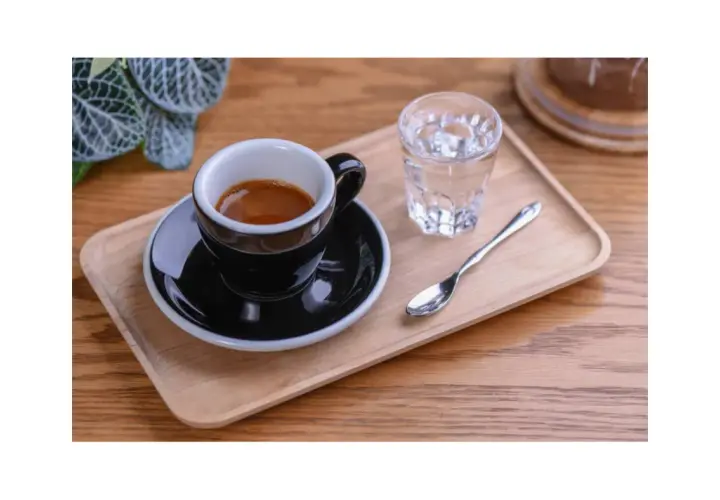 Czarna filiżanka espresso na drewnianej tacy, obok szklanka wody i łyżeczka. Idealnie pokazuje, jak podawać espresso.