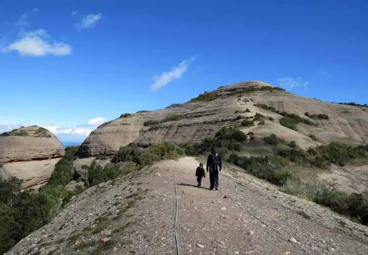Un padre y su hijo caminan por un sendero rocoso hacia la cima del Montcau, bajo un cielo azul con nubes dispersas.