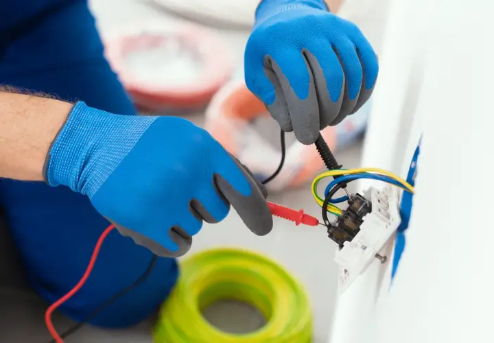 Elektryk w niebieskich rękawicach sprawdza połączenia przewodów. W tle widać rolki kabli. To instrukcja, jak podłączyć gniazdko bez uziemienia.