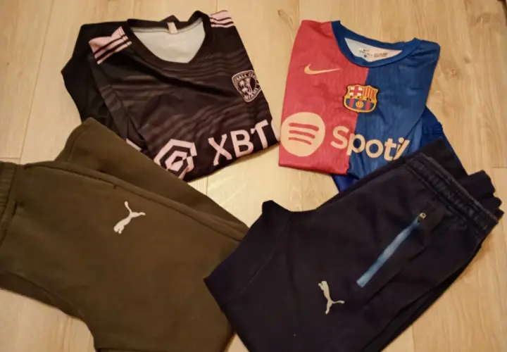 Dwa komplety odzieży piłkarskiej dla chłopca: czarna koszulka z różowymi pasami i herbem, niebiesko-czerwona koszulka FC Barcelony, brązowe i granatowe spodnie piłkarskie chłopięce z logo Pumy.