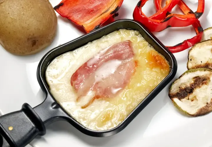 Un poêlon de raclette avec fromage fondu et lardons, accompagné d'une pomme de terre, de poivrons rouges grillés et de courgettes. Une recette de sauce raclette facile et gourmande.