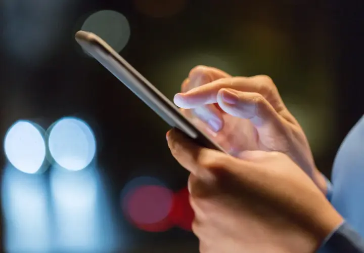 Dłoń trzyma smartfon, palec wskazujący dotyka ekranu, być może w trakcie odzyskiwania zdjęć z telefonu. Rozmyte światła w tle sugerują noc.