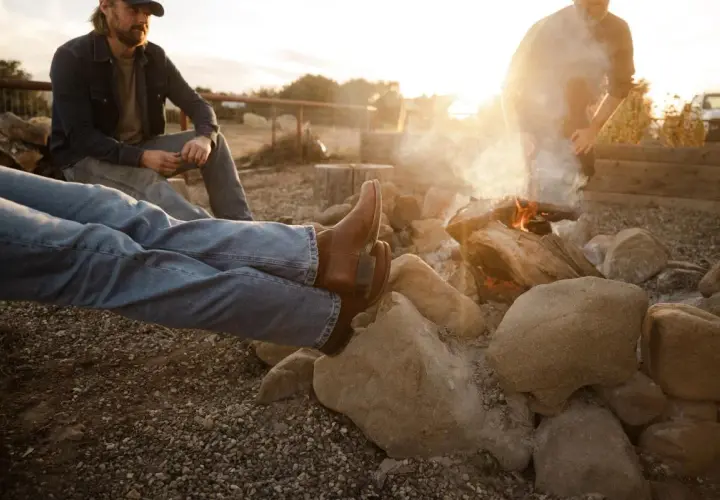 Two men relax by a campfire. One wears brown boots, and the other, a cap. The sun sets, casting a warm glow.