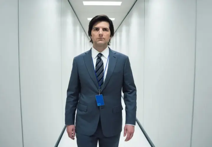 Uomo in abito grigio cammina in un corridoio bianco, con un badge blu appeso al collo. La sua espressione è seria, quasi a suggerire una scissione nella sua recensione.