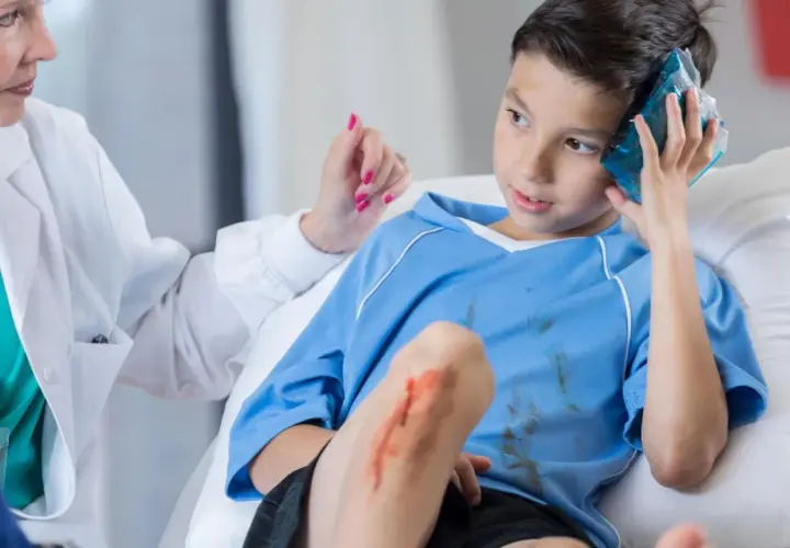 Niño con golpe en la cabeza, recibe atención médica. Tiene una compresa fría en la sien y una herida en la rodilla.