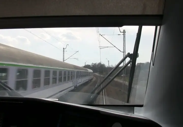 Widok z kabiny maszynisty na mijający pociąg. Widać tory, sieć trakcyjną i niebo z chmurami.